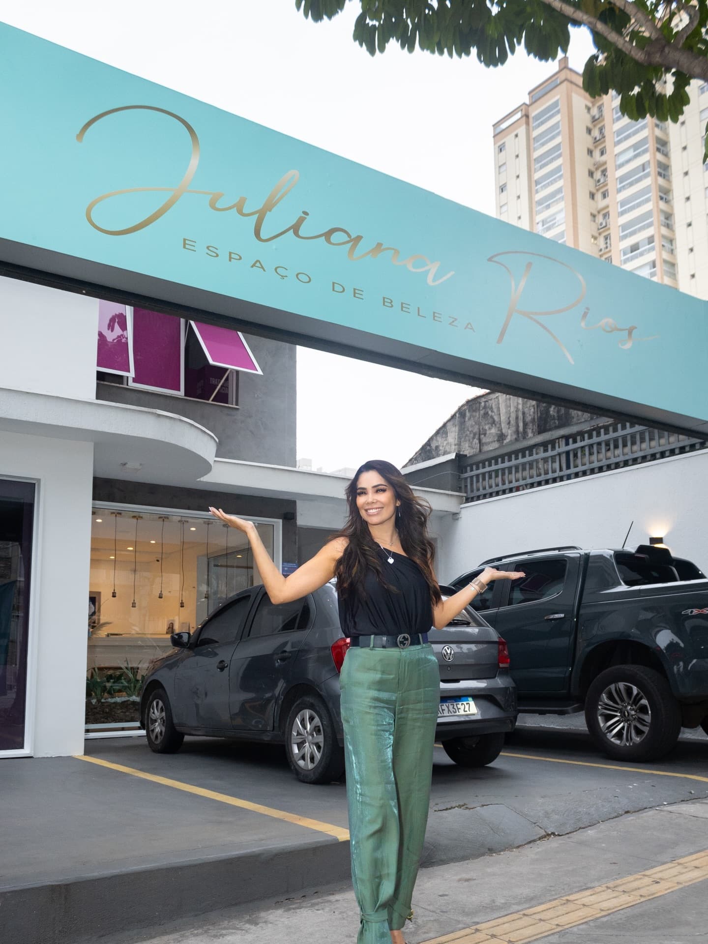 Juliana Rios de braços abertos na porta do espaço, embaixo do letreiro turquesa